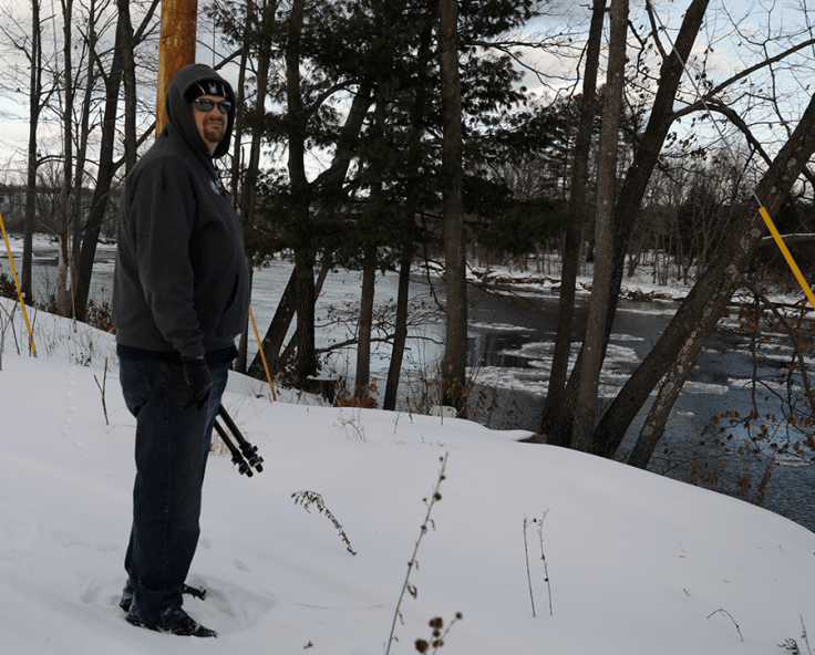 Man holding a tripod overlooking a partially frozen river